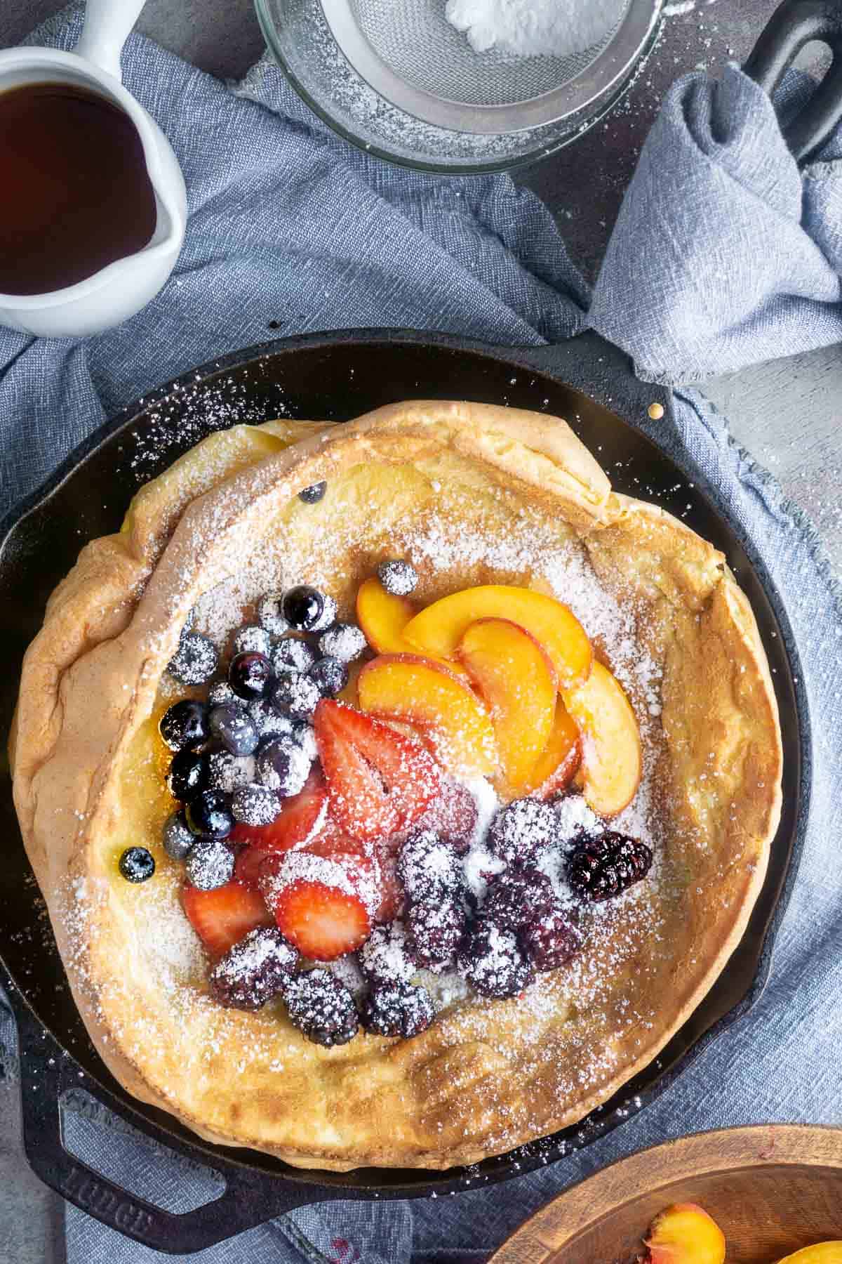 Fruit topped Dutch baby pancake in iron skillet next to maple syrup and powdered sugar