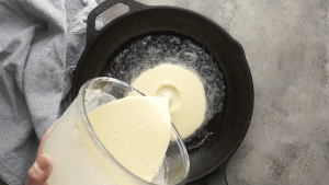 Dutch baby batter pouring into iron skillet next to gray linen on gray surface