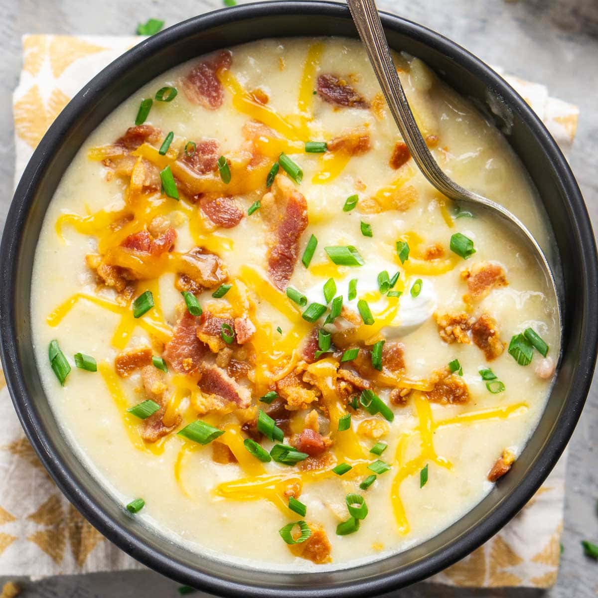 A bowl of loaded potato soup topped with shredded cheddar cheese, chopped bacon, and green onions, served with a spoon in the bowl.