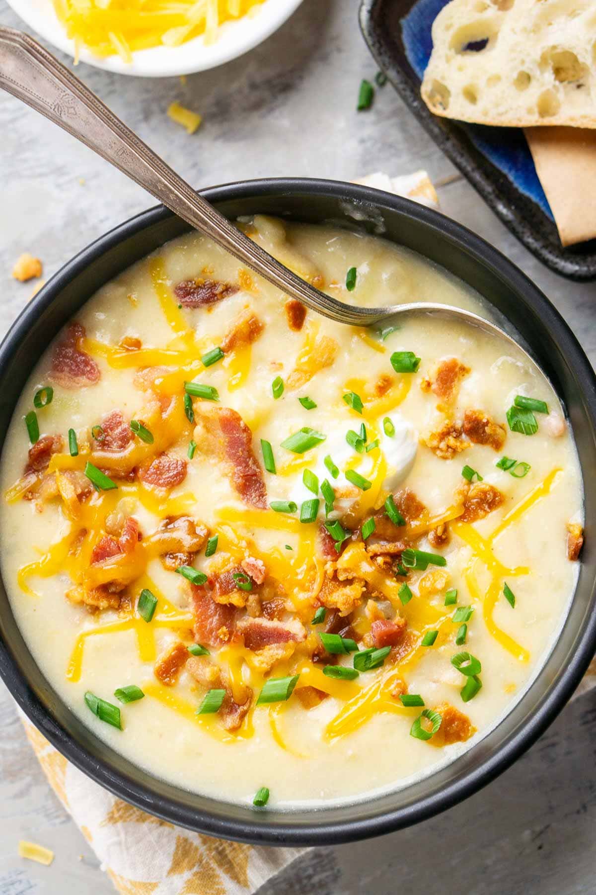 A bowl of creamy loaded potato soup topped with shredded cheese, bacon bits, and chopped chives, served with a spoon in the bowl and bread on the side.