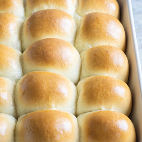 A white baking dish filled with golden brown, freshly baked dinner rolls closely arranged together.
