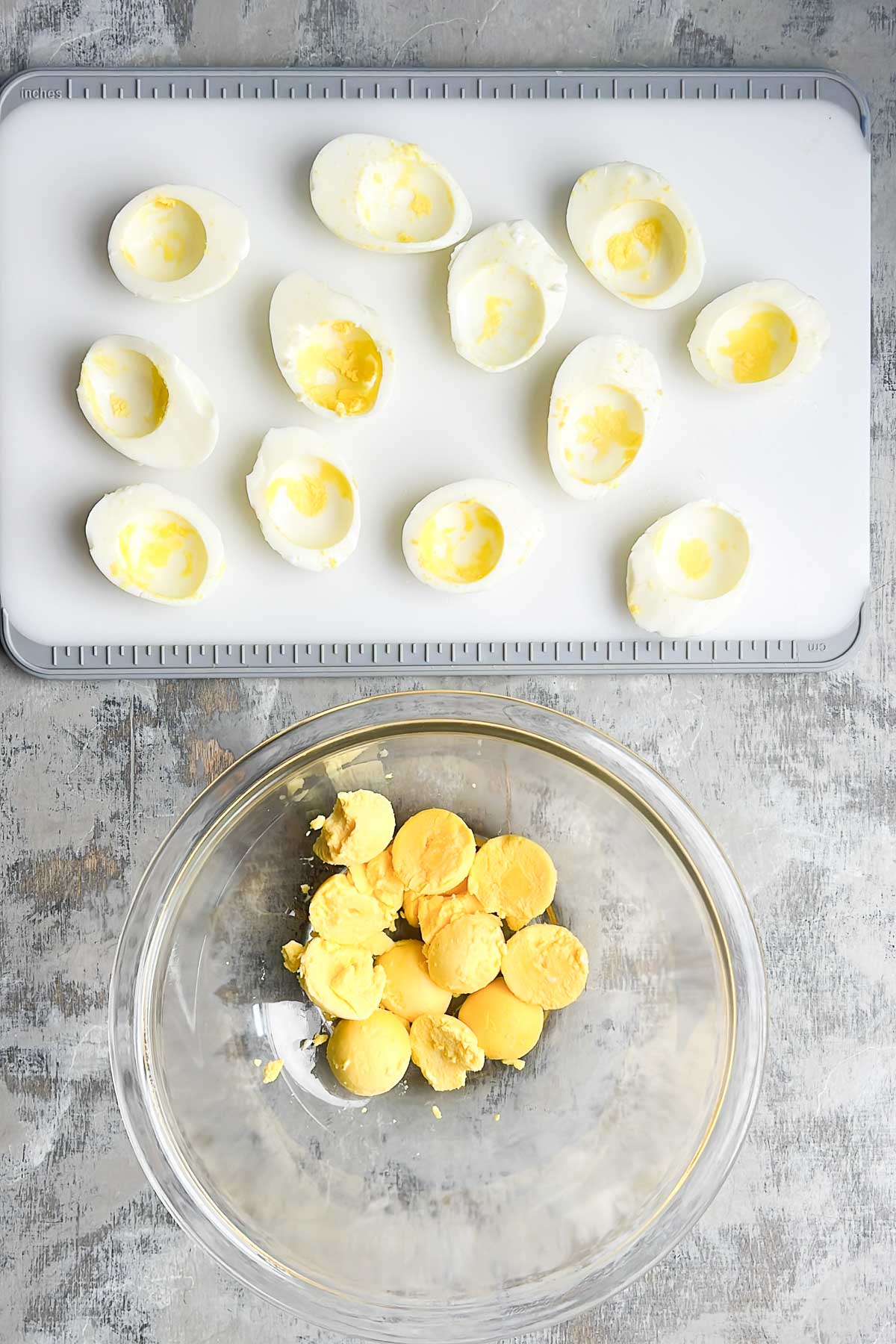A cutting board with empty hard-boiled egg white halves and a clear bowl containing cooked egg yolks on a gray surface.