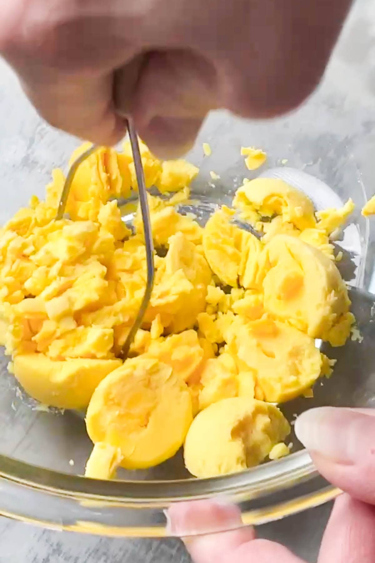 A hand uses a metal masher to mash hard-boiled egg yolks in a clear glass bowl.