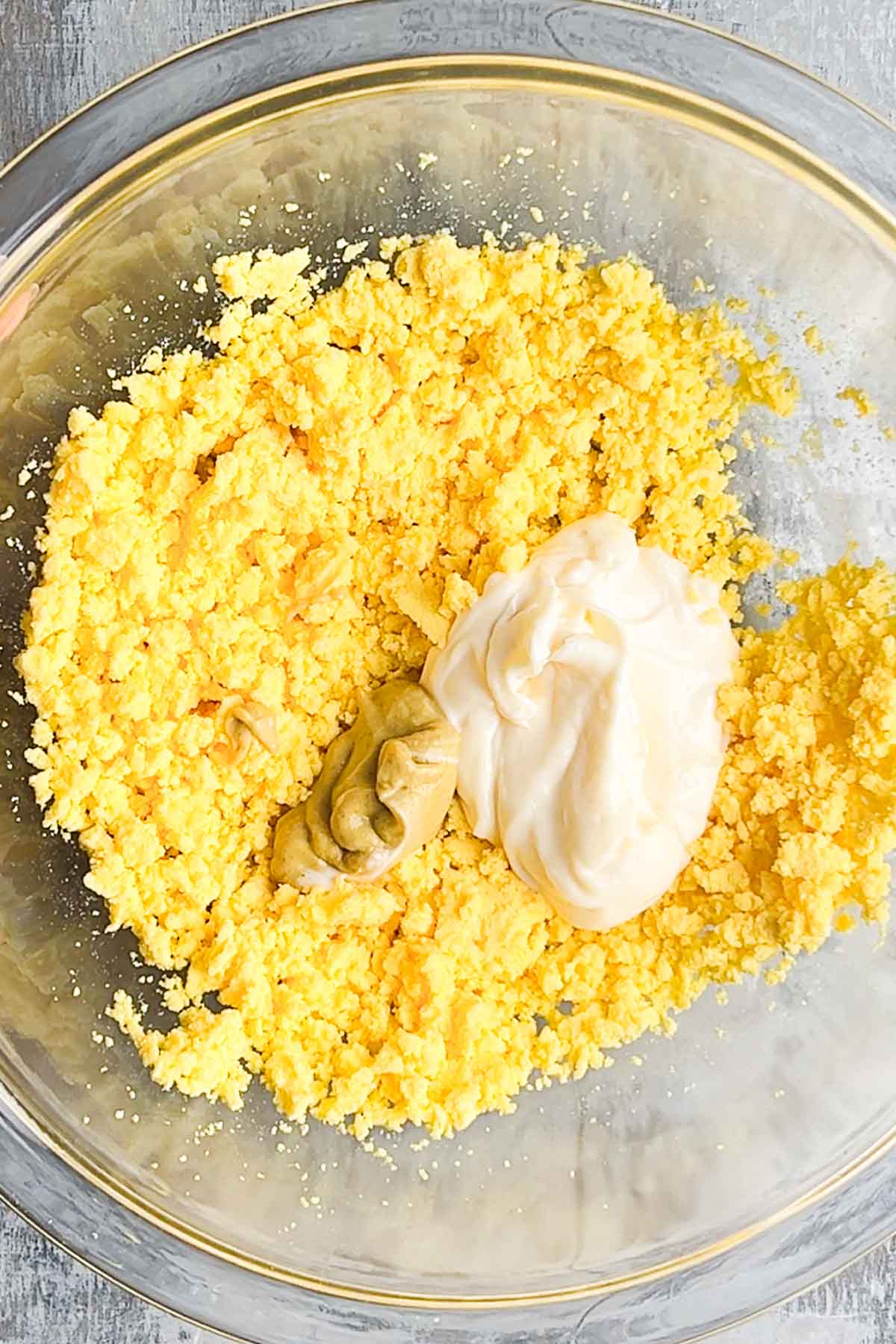 A glass bowl containing crumbled cooked egg yolks with dollops of mayonnaise and mustard on top.
