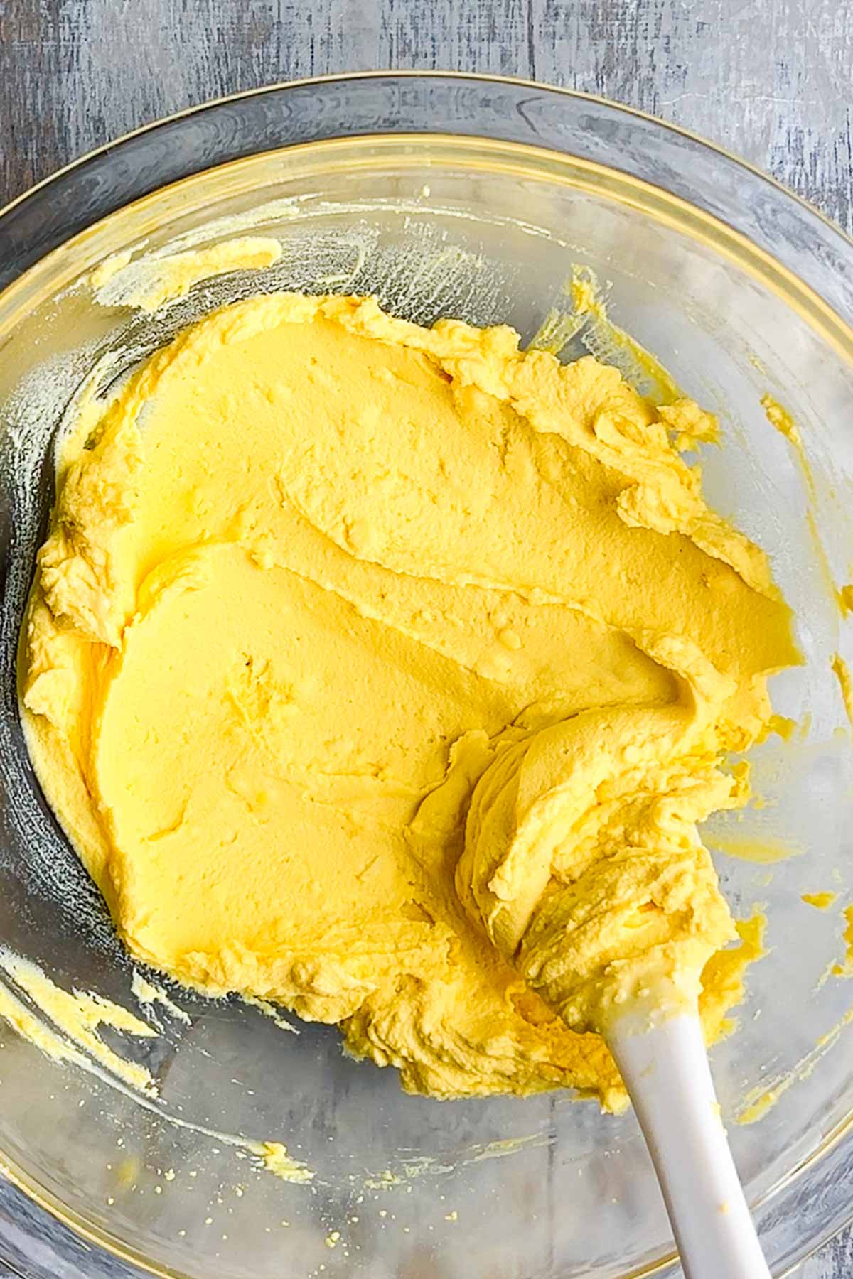 A glass bowl containing smooth yellow egg salad mixture with a white spatula on a gray surface.