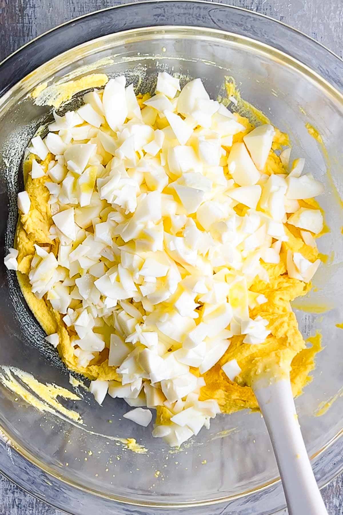A glass bowl containing chopped hard-boiled eggs on top of a yellow egg salad mixture, with a white spatula partially visible.