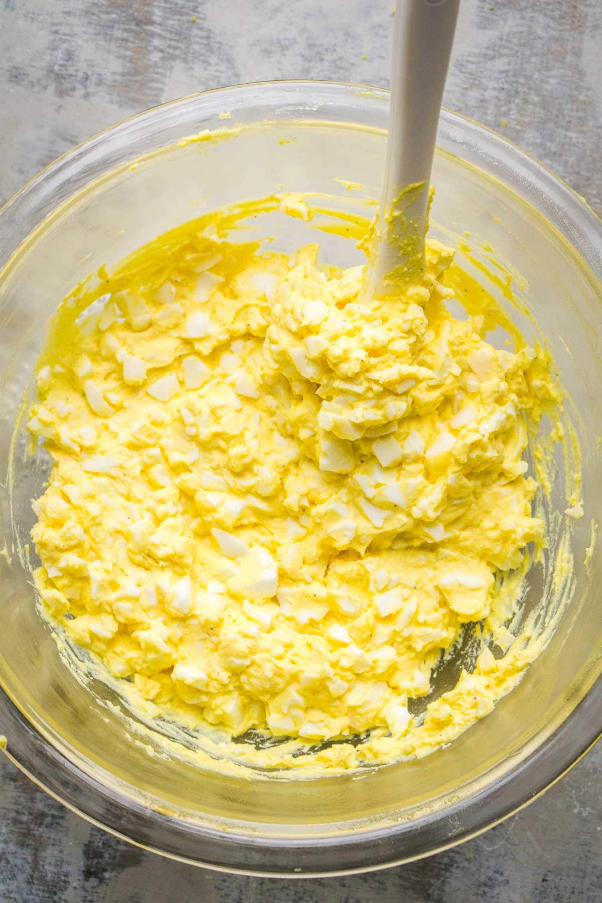A glass bowl with egg salad being stirred with a white spatula.