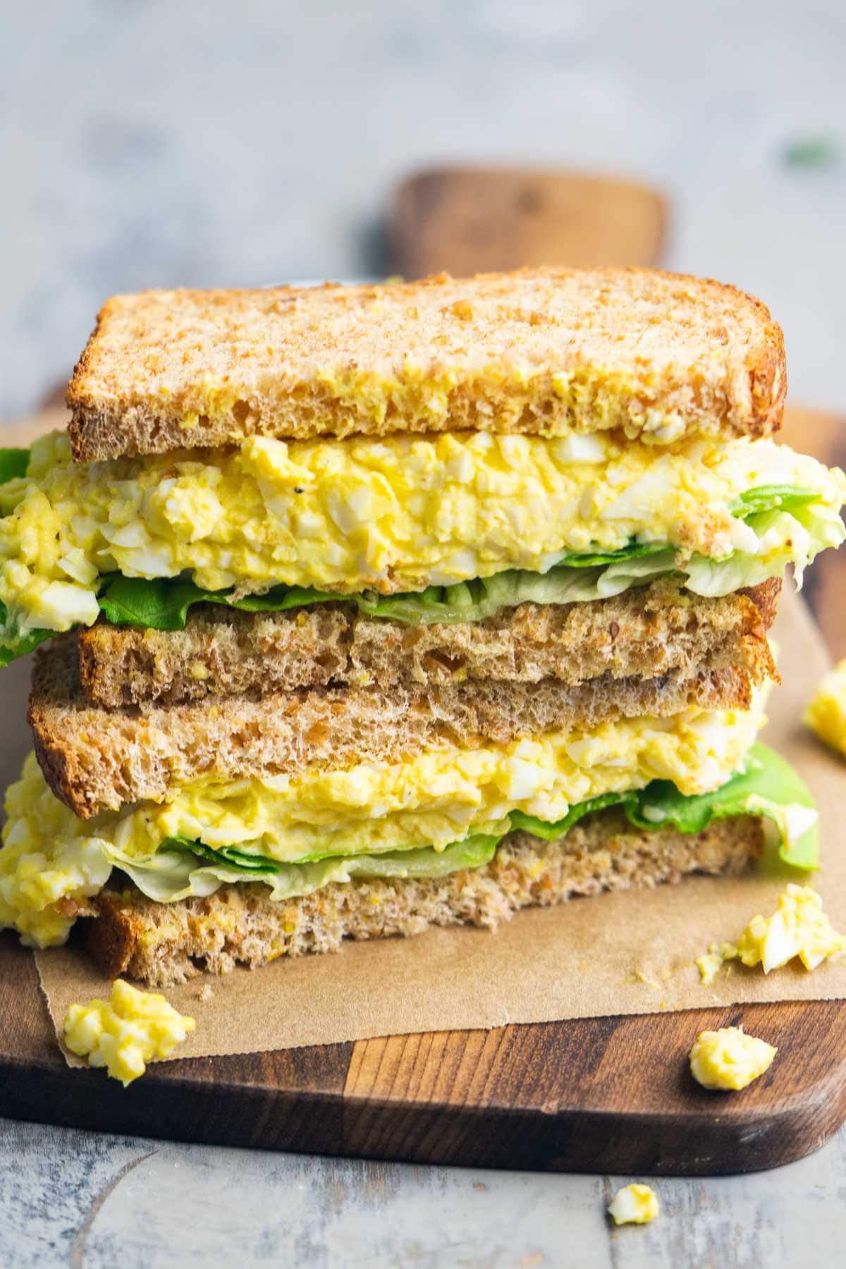 A classic egg salad sandwich with leafy greens, stacked on whole wheat bread, cut in half and served on parchment paper atop a wooden board.