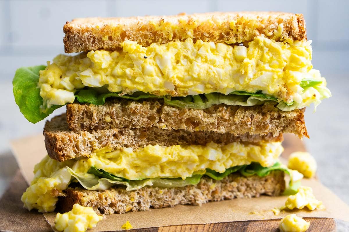 A classic egg salad sandwich with leafy greens, stacked on whole wheat bread, cut in half and served on parchment paper atop a wooden board.
