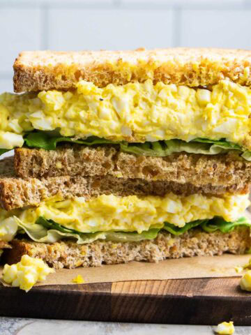 A classic egg salad sandwich with leafy greens, stacked on whole wheat bread, cut in half and served on parchment paper atop a wooden board.