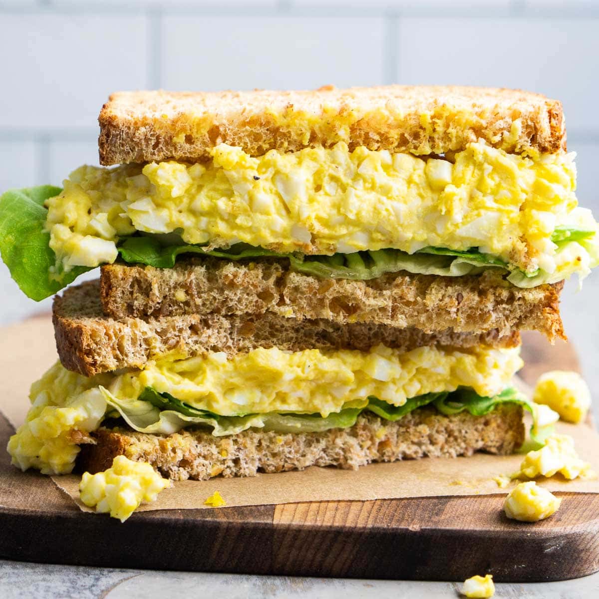 A classic egg salad sandwich with leafy greens, stacked on whole wheat bread, cut in half and served on parchment paper atop a wooden board.