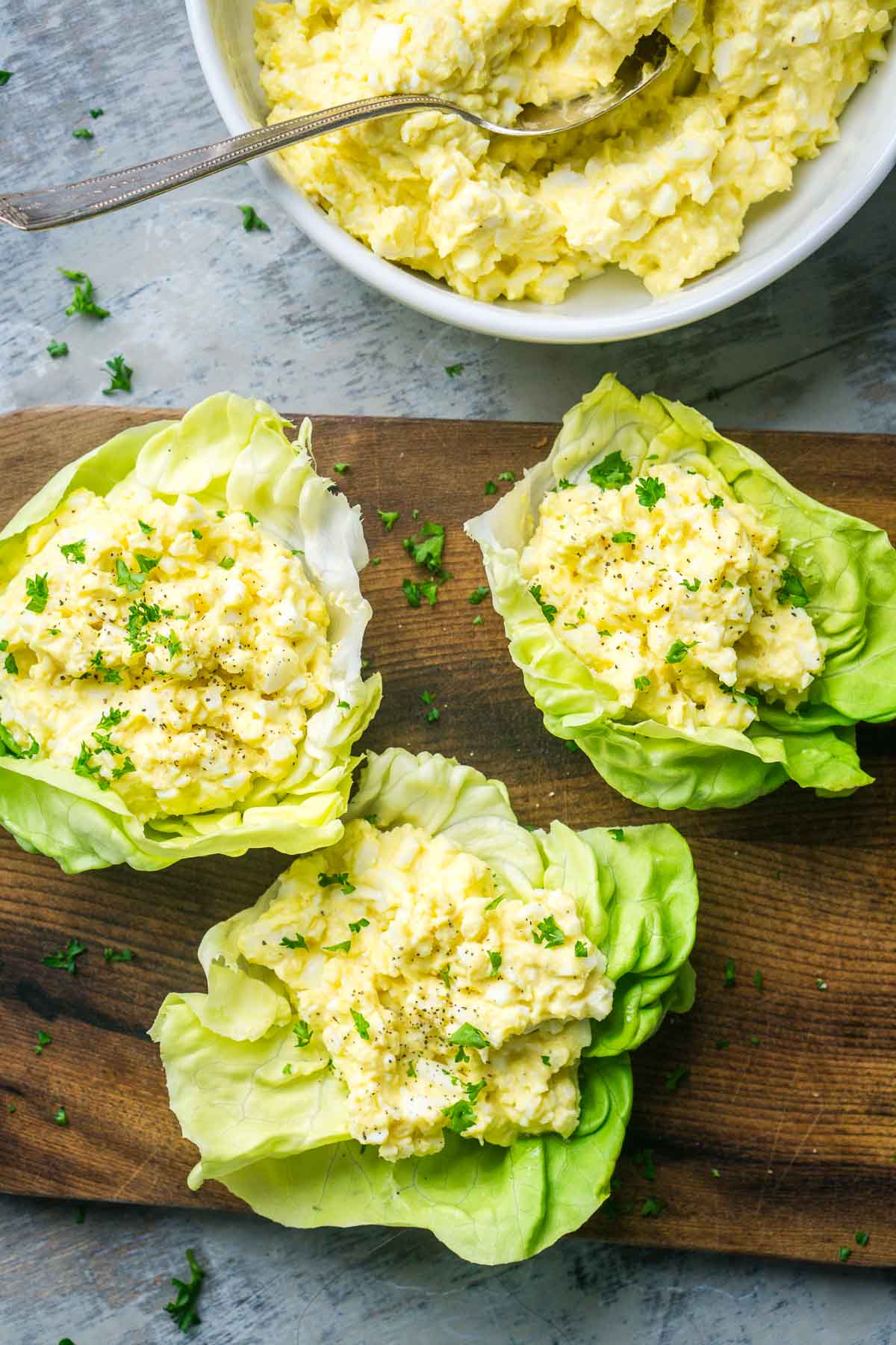 Three egg salad lettuce wraps arranged on a wooden board garnished with fresh parsley, with a bowl of extra egg salad and a spoon in the background.