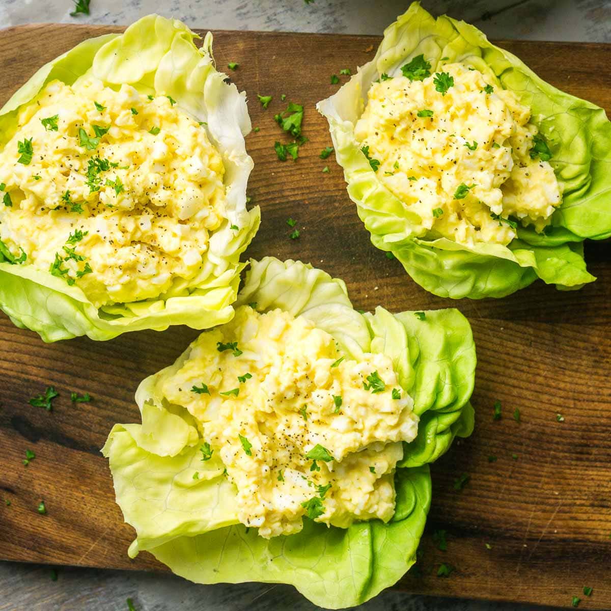 Three egg salad lettuce wraps arranged on a wooden board garnished with fresh parsley, with a bowl of extra egg salad and a spoon in the background.