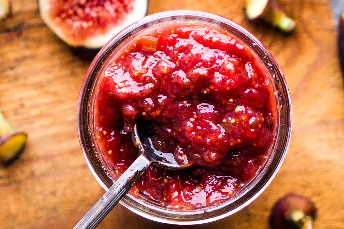 A glass jar filled with chunky red fig jam, with a spoon inside, sits on a wooden surface surrounded by fresh fig pieces.