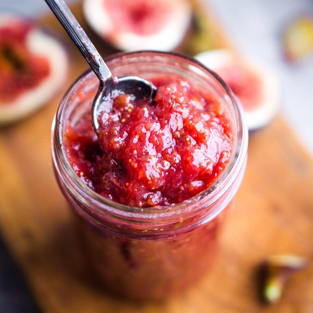 A glass jar filled with chunky red fig jam, with a spoon inside, sits on a wooden surface surrounded by fresh fig pieces.