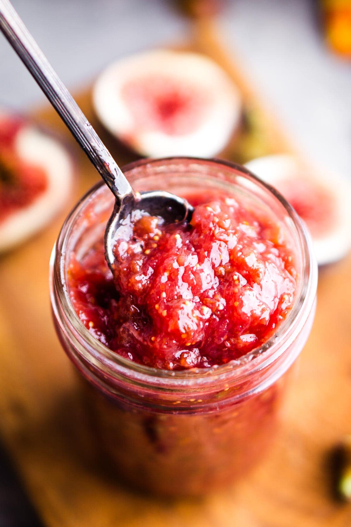 A glass jar filled with chunky red fig jam, with a spoon inside, sits on a wooden surface surrounded by fresh fig pieces.