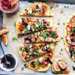 Flatbread topped with figs, prosciutto, arugula, goat cheese, walnuts, and balsamic glaze, sliced on parchment paper with fresh figs and sauce on the side.