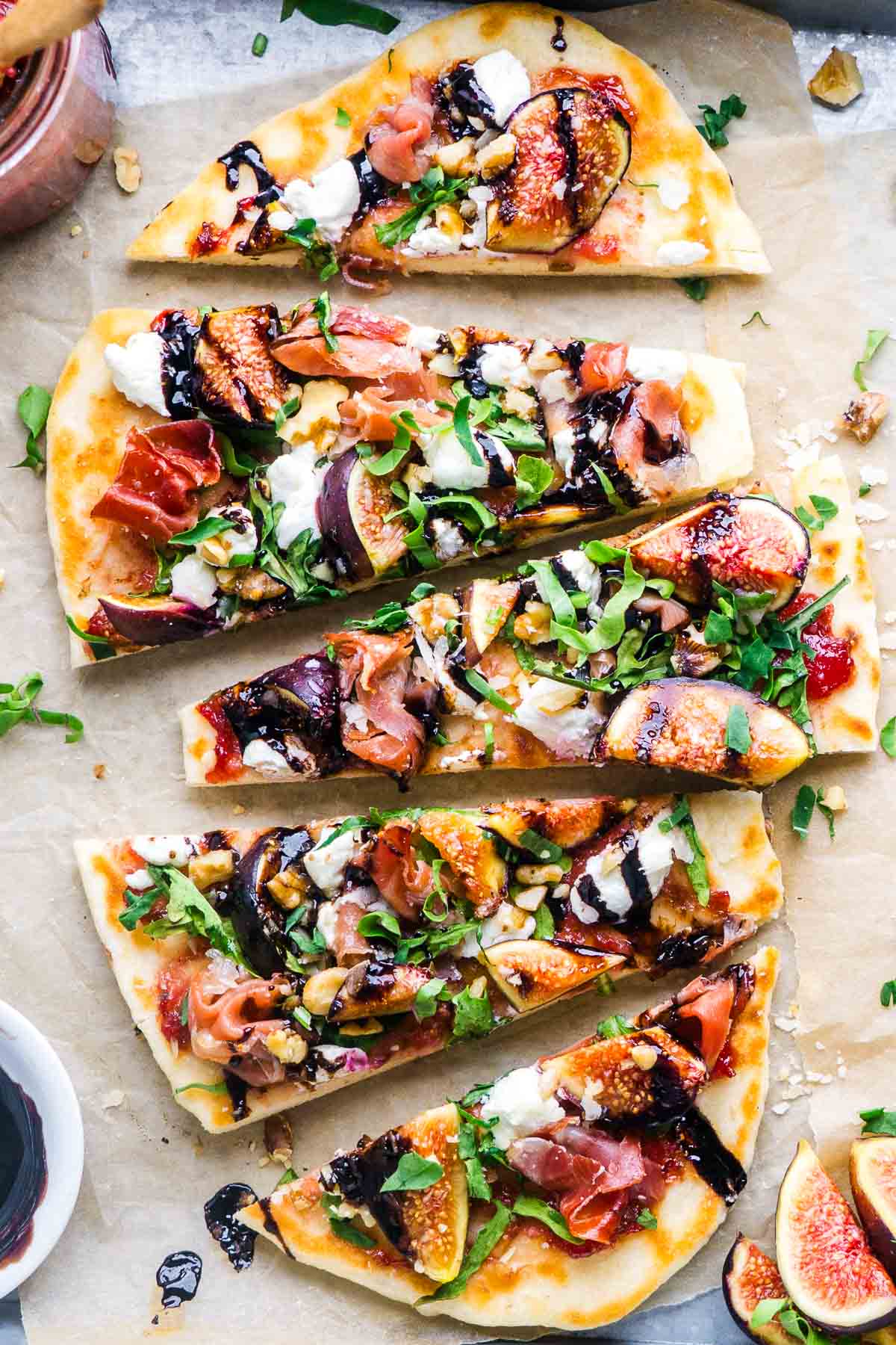 Flatbread topped with figs, prosciutto, arugula, goat cheese, walnuts, and balsamic glaze, sliced on parchment paper with fresh figs and sauce on the side.