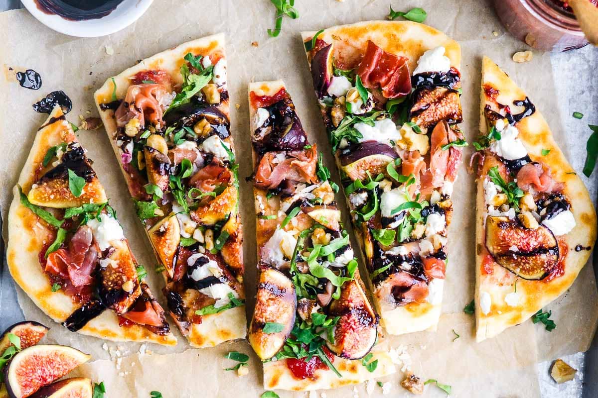 Flatbread topped with figs, prosciutto, arugula, goat cheese, walnuts, and balsamic glaze, sliced on parchment paper with fresh figs and sauce on the side.