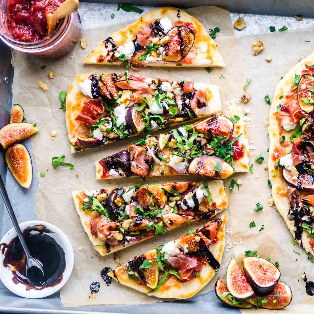 Flatbread topped with figs, prosciutto, arugula, goat cheese, walnuts, and balsamic glaze, sliced on parchment paper with fresh figs and sauce on the side.