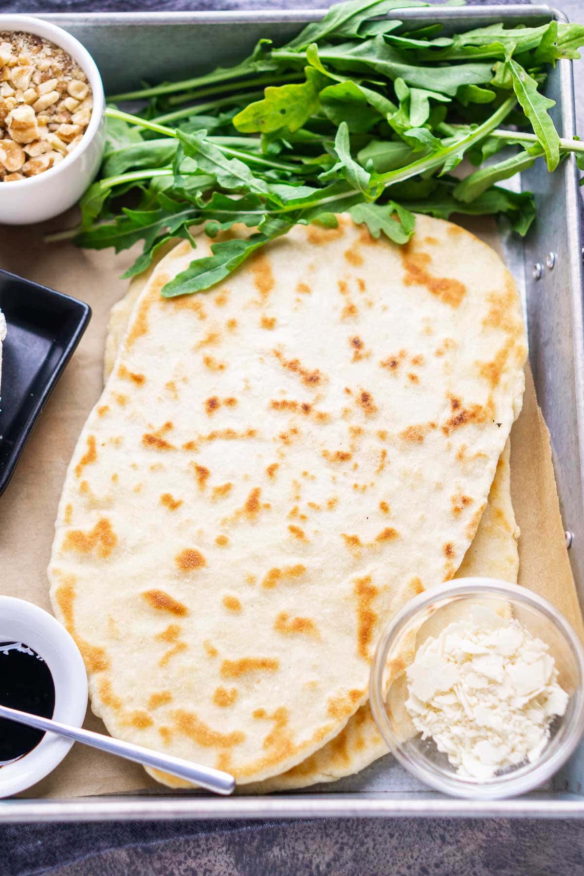 Two pieces of flatbread on parchment paper, with fresh arugula, a bowl of walnuts, crumbled cheese, and a small dish of dark sauce beside them.