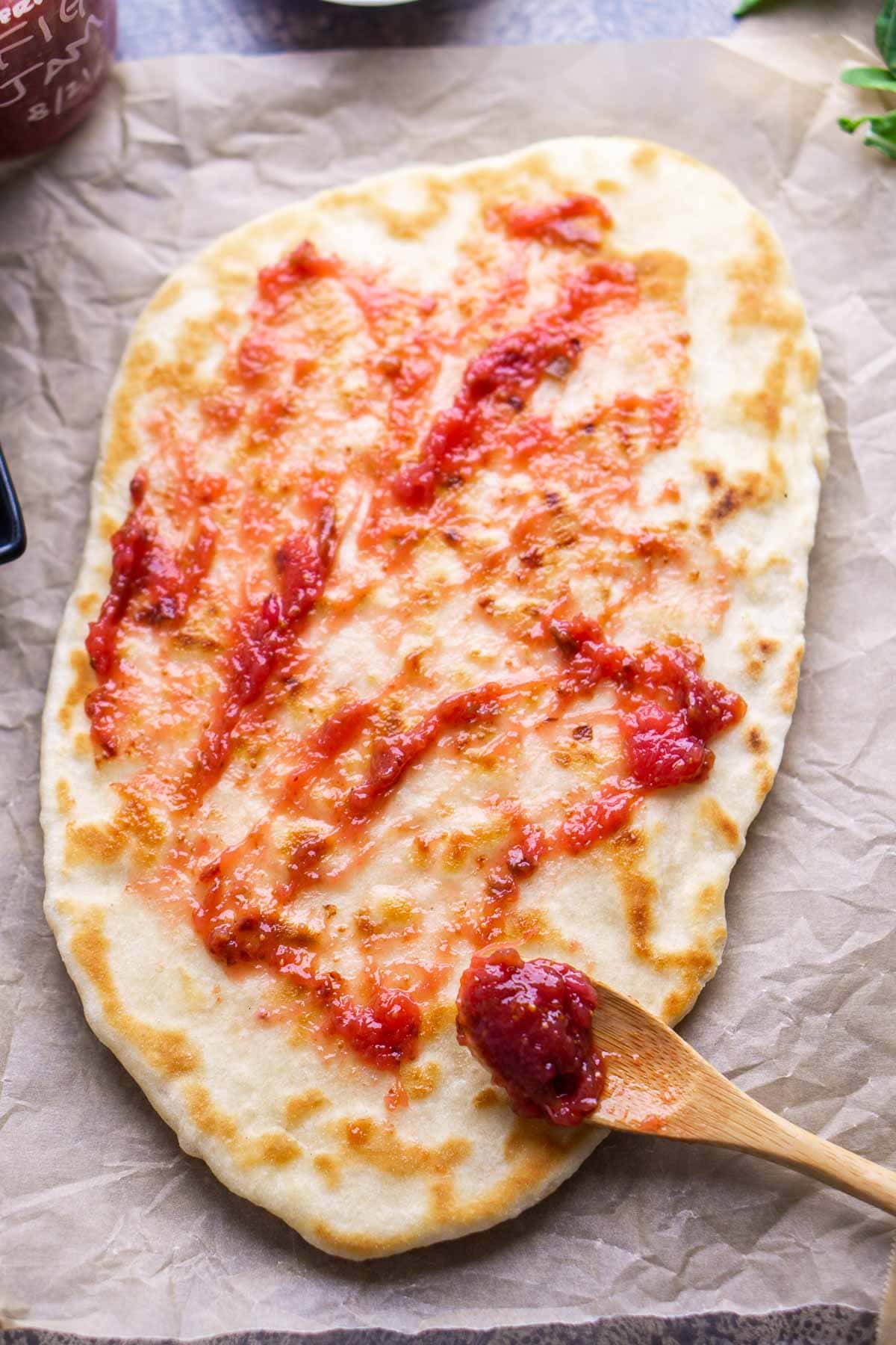 Flatbread on parchment paper being spread with fig jam using a wooden spoon.