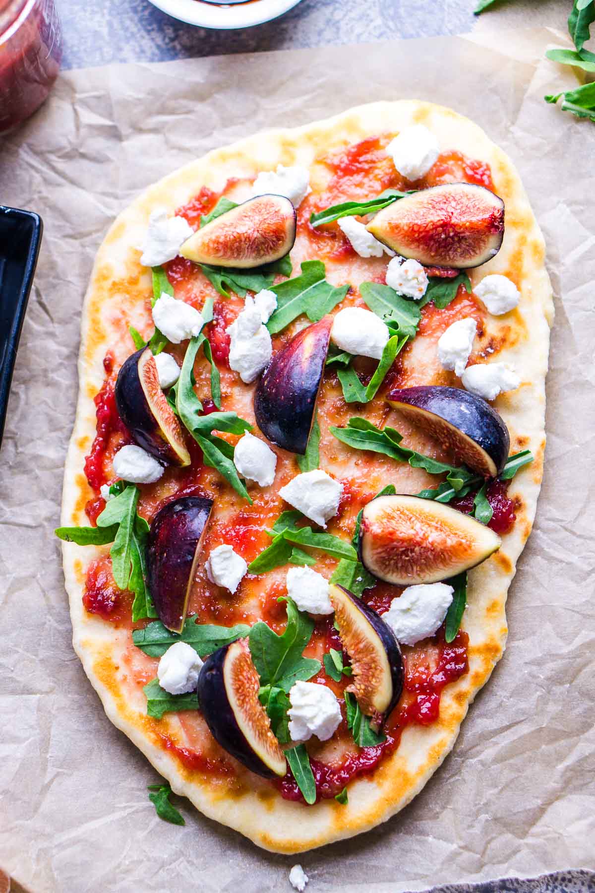 Oval-shaped flatbread topped with fig jam, arugula, fresh figs, and dollops of goat cheese, served on parchment paper.