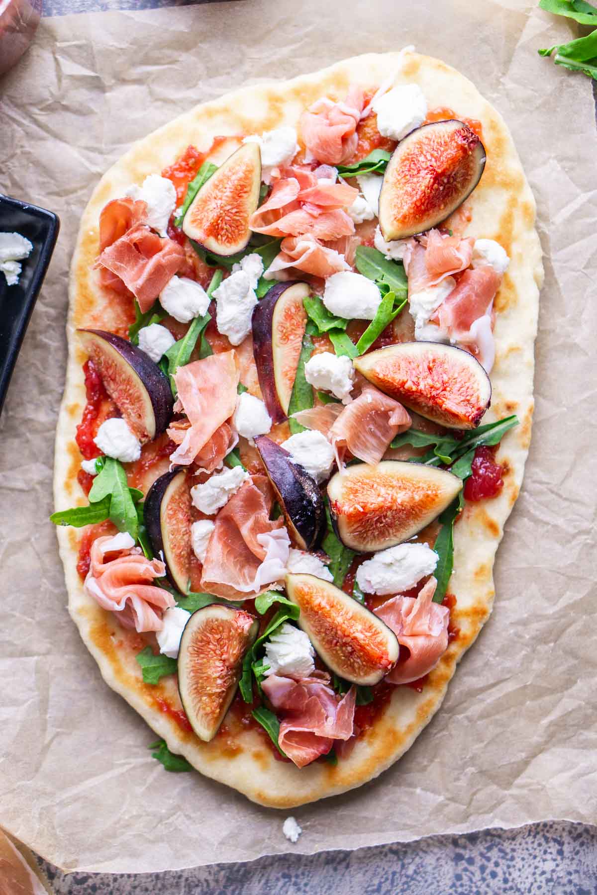 Oval-shaped flatbread topped with fig jam, arugula, fresh figs, prosciutto, and dollops of goat cheese, served on parchment paper.