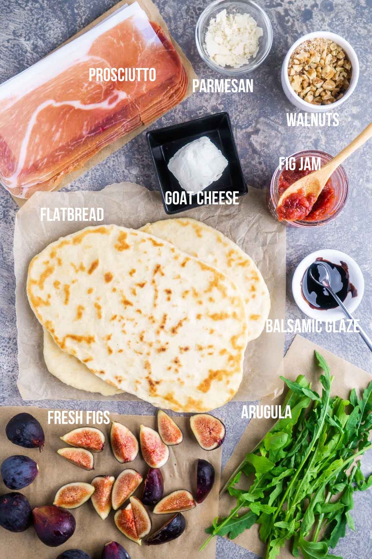 Flatbread surrounded by ingredients labeled as prosciutto, parmesan, walnuts, goat cheese, fig jam, balsamic glaze, fresh figs, and arugula, arranged on a gray surface.