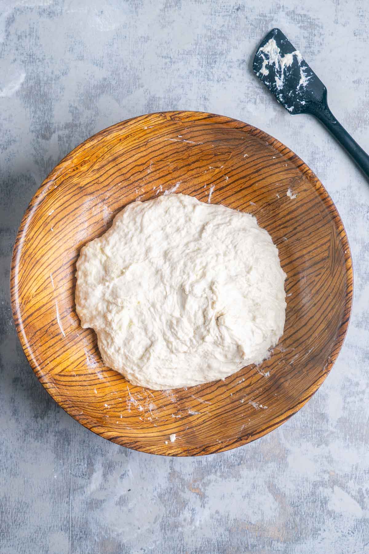 A wooden bowl containing a ball of freshly mixed raw bread dough sits on a gray surface next to a black spatula with some dough residue.