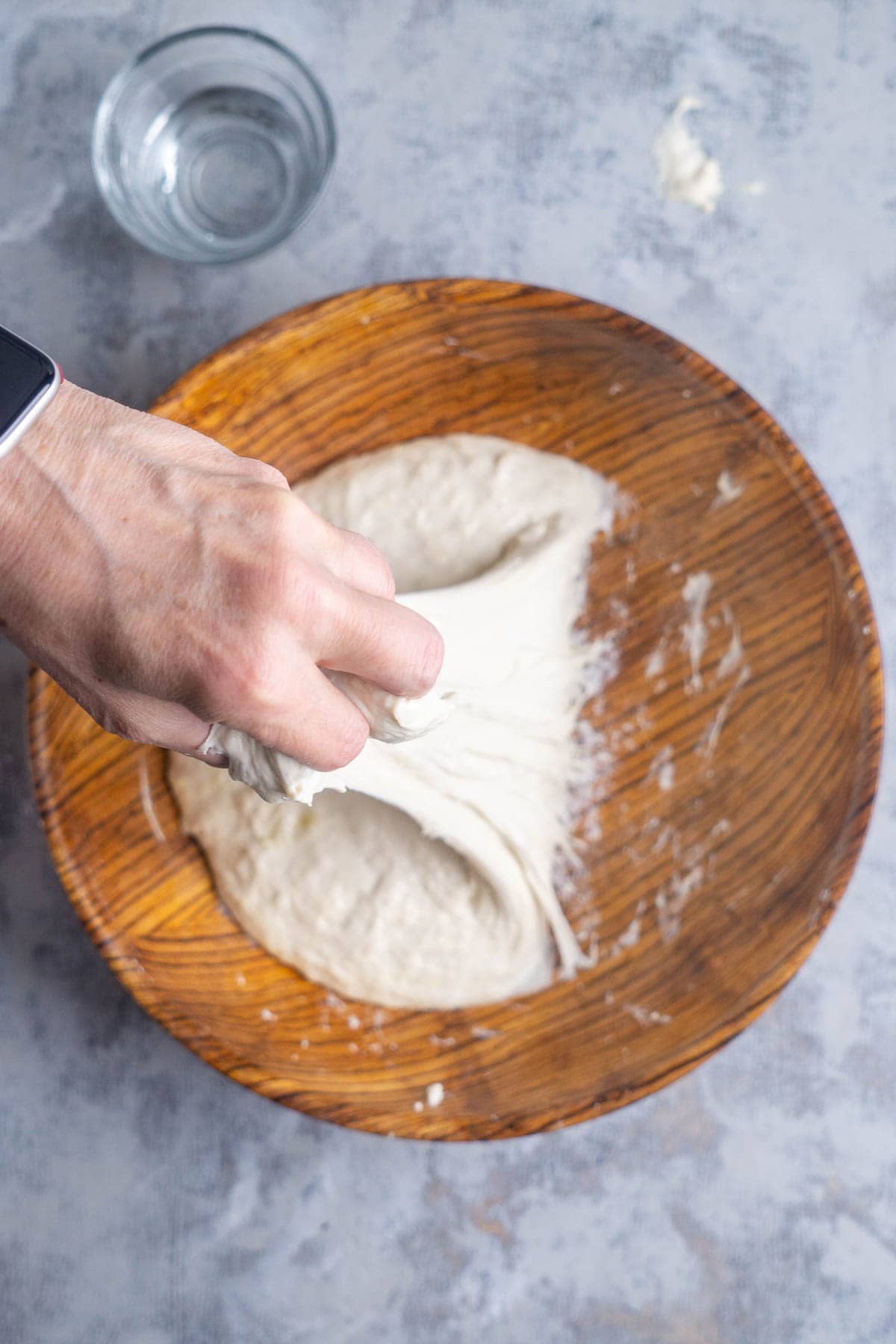 A hand stretches bread dough in a wooden bowl, with a glass of water placed nearby on a gray surface.