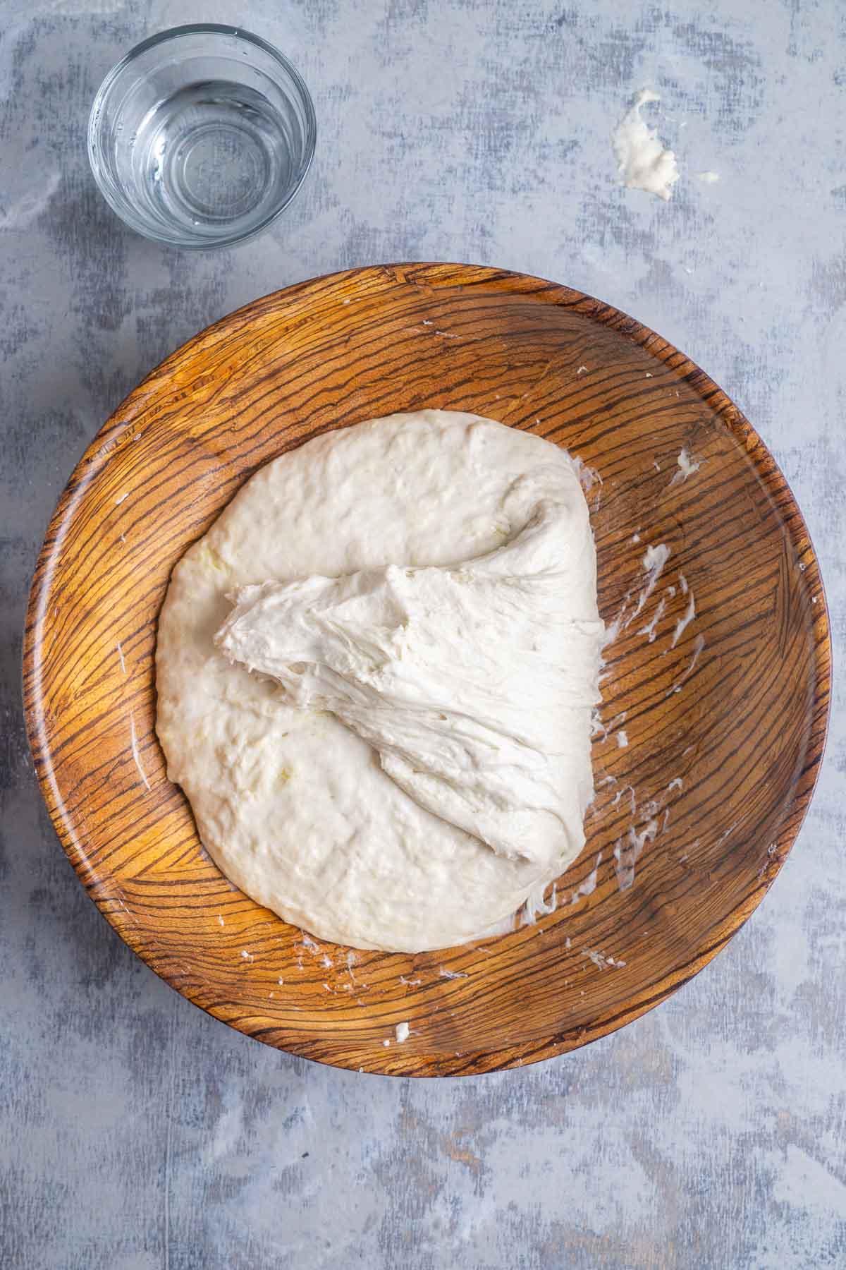 A wooden bowl containing bread dough next to a glass of water on a lightly floured surface.
