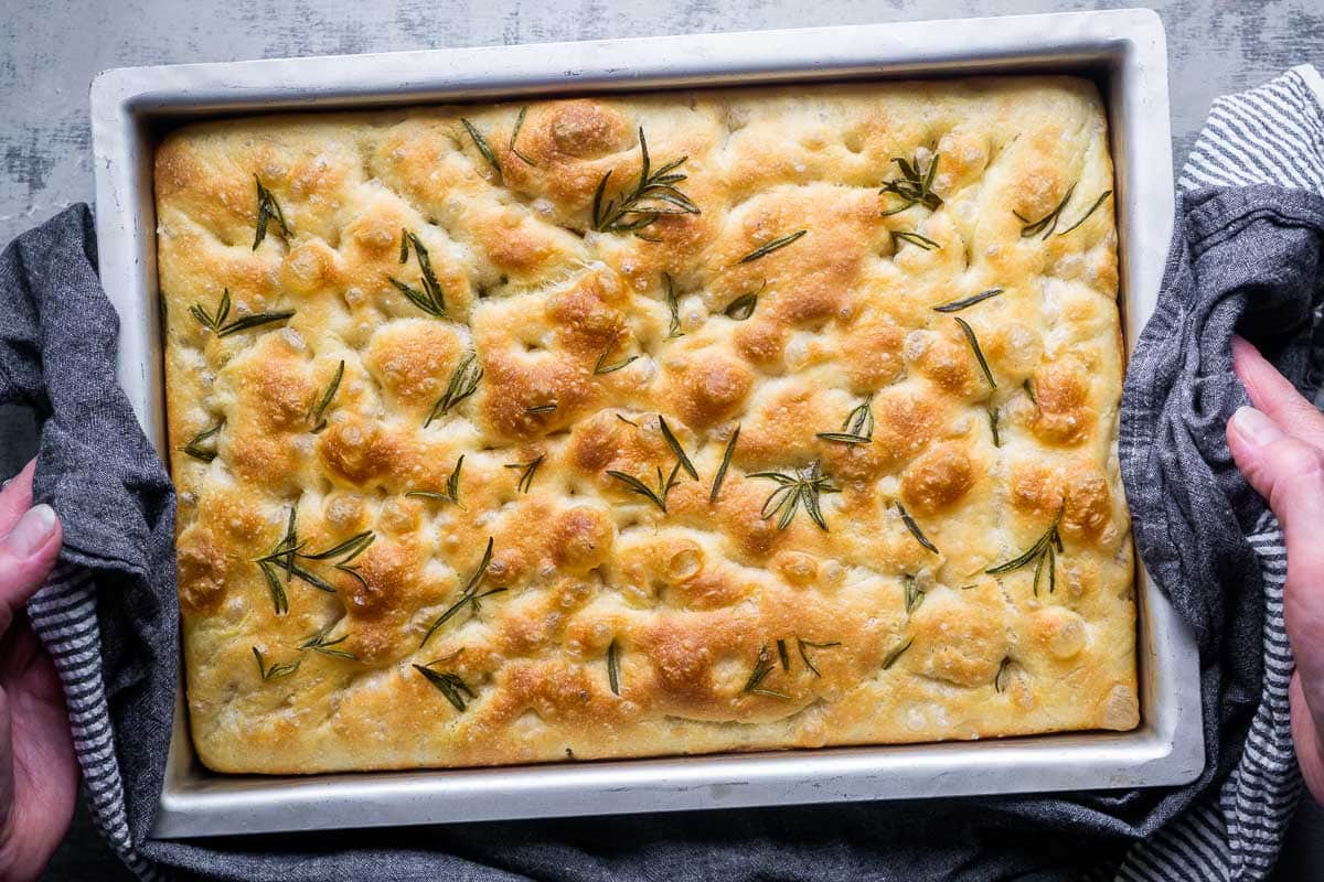 A rectangular pan of golden-brown focaccia bread, fragrant with rosemary sprigs, is held by hands using a dark cloth.