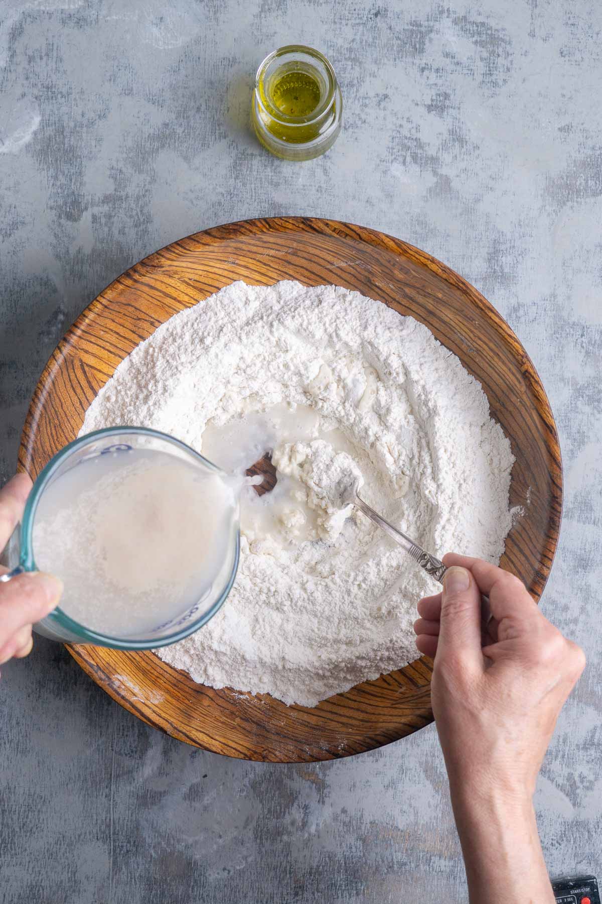 A person pours a yeast and water mixture into a well of flour in a wooden bowl, with a small jar of oil nearby on a gray surface.