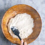 Hand mixing focaccia bread dough with a black spatula in a wooden bowl on a gray textured surface.