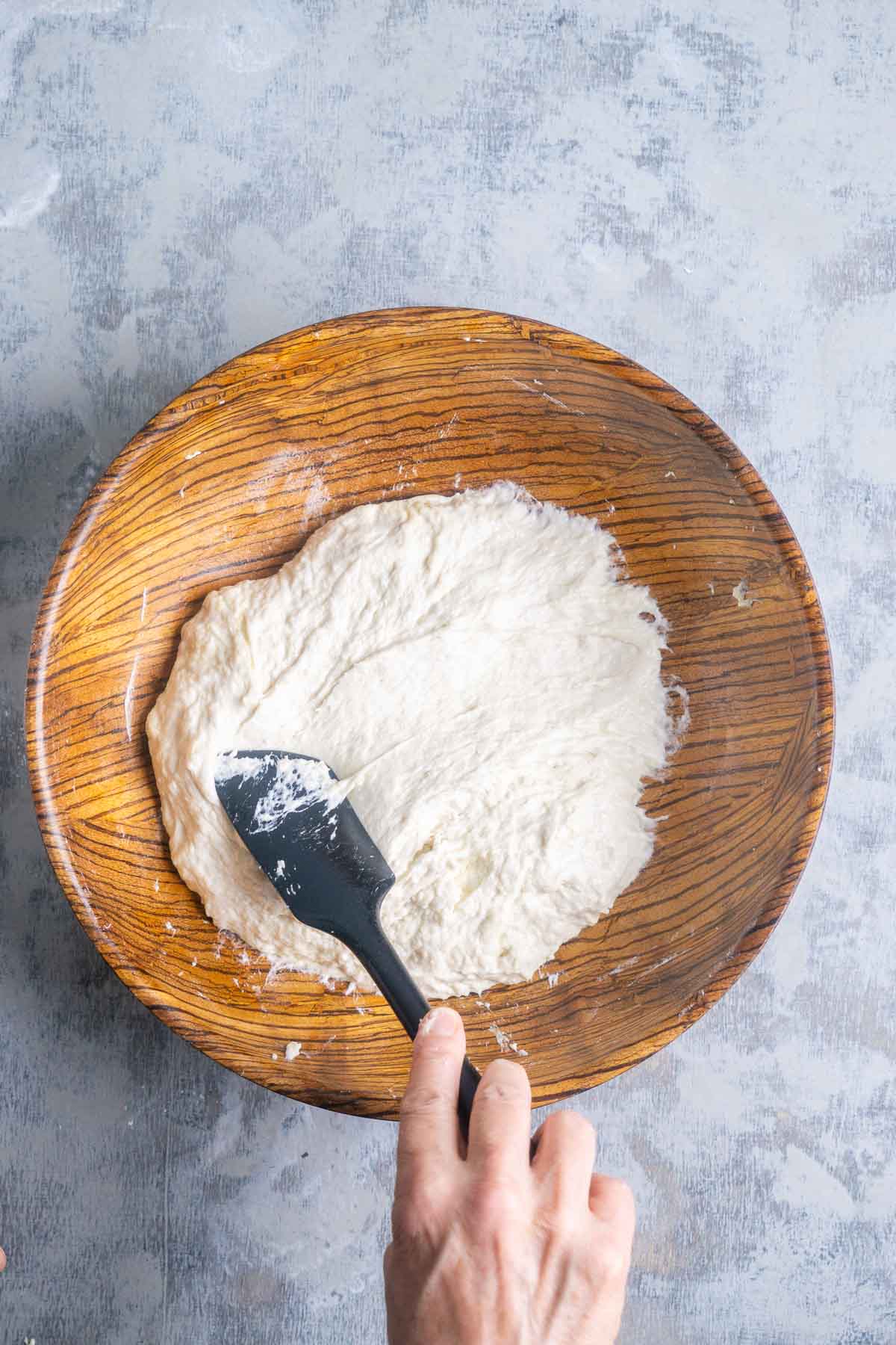 Hand mixing focaccia bread dough with a black spatula in a wooden bowl on a gray textured surface.