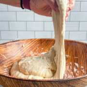 A hand stretches sticky focaccia bread dough in a wooden bowl against a white tile background.
