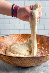 A hand stretches sticky focaccia bread dough in a wooden bowl against a white tile background.
