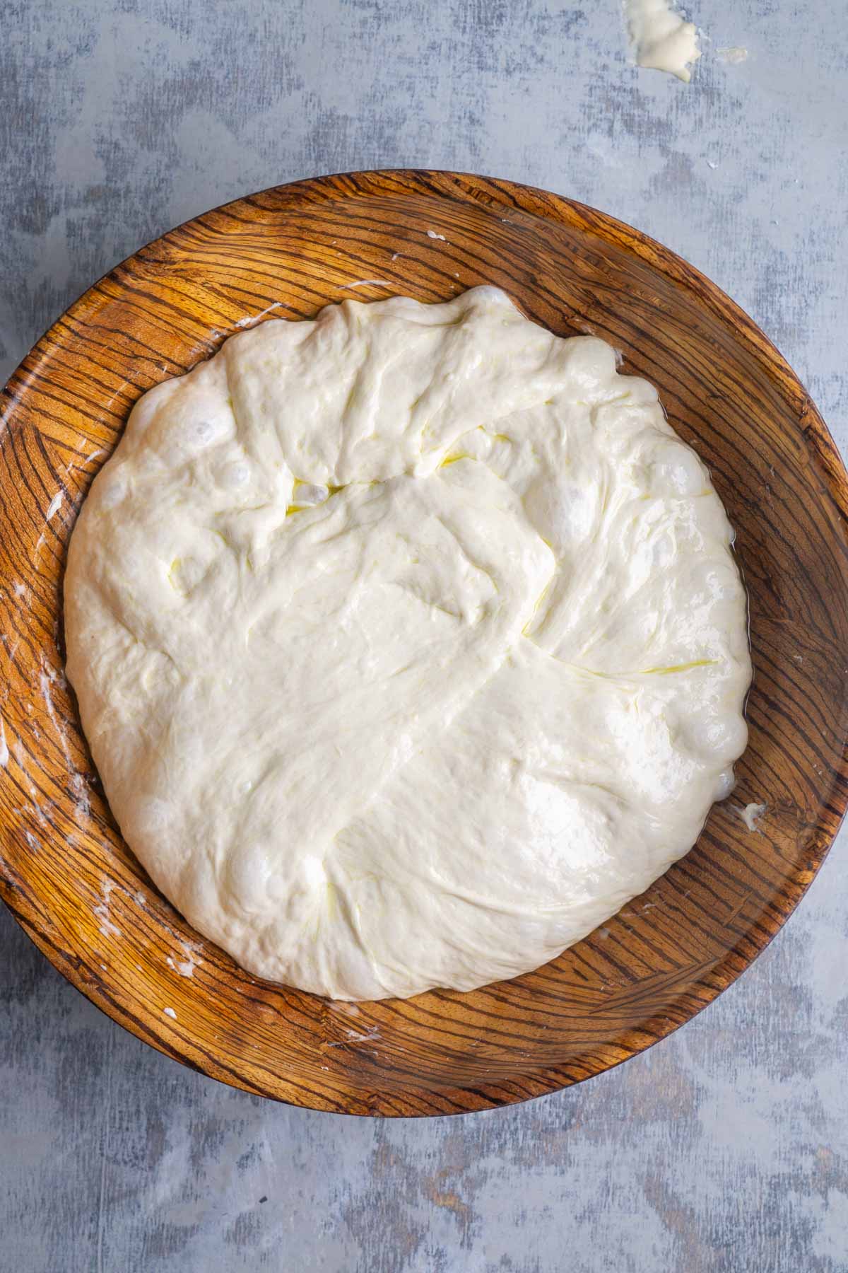 A wooden bowl filled with bubbly focaccia bread dough sits on a light gray countertop.