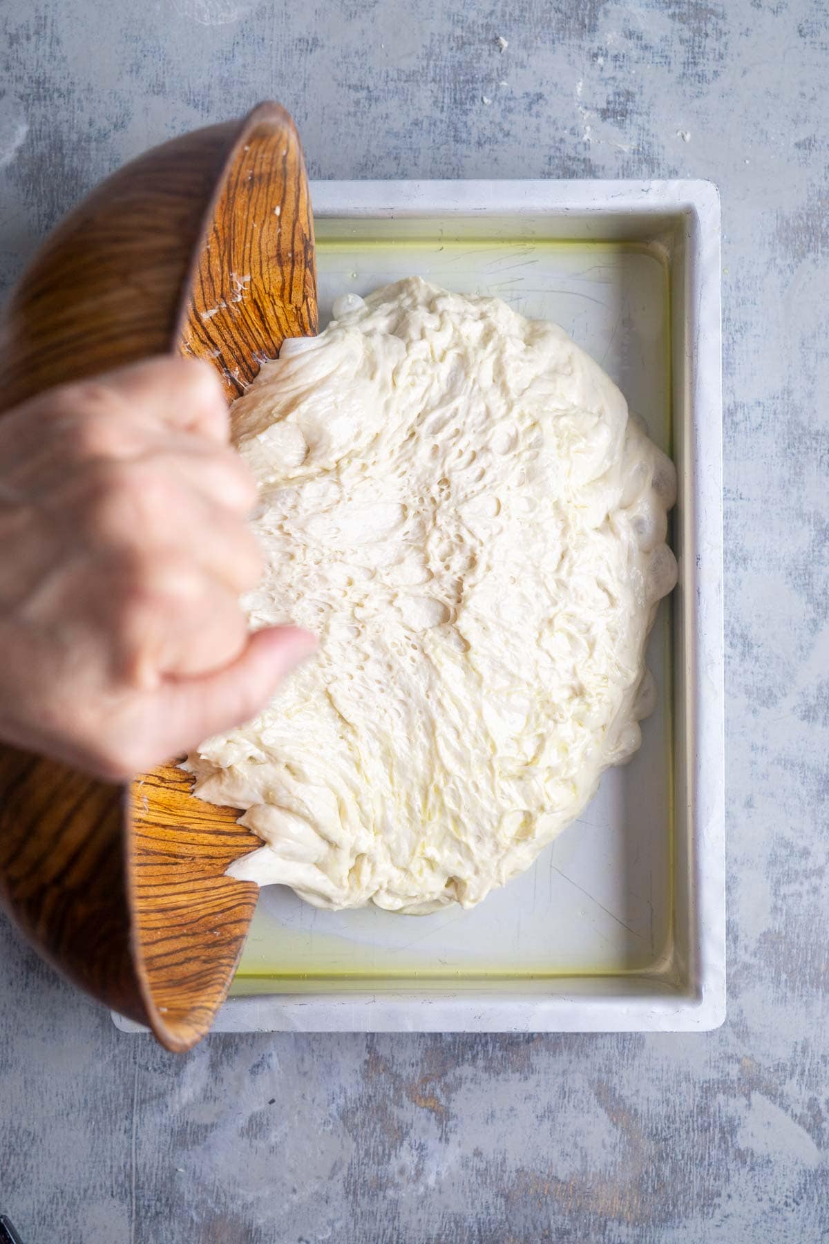 A hand is transferring puffy focaccia dough from a wooden bowl into a rectangular baking pan with a light coating of oil.