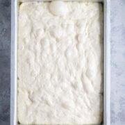 A rectangular baking pan filled with raw, bubbly focaccia bread dough rests on a light textured surface.