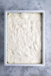 A rectangular baking pan filled with raw, bubbly focaccia bread dough rests on a light textured surface.