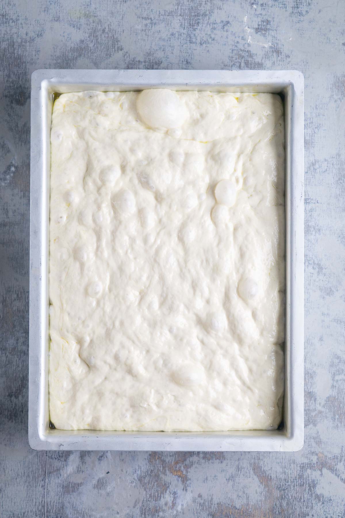A rectangular baking pan filled with raw, bubbly focaccia bread dough rests on a light textured surface.
