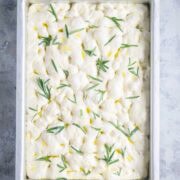 A rectangle pan filled with raw focaccia bread dough topped with rosemary sprigs and drizzled with olive oil, ready to be baked.