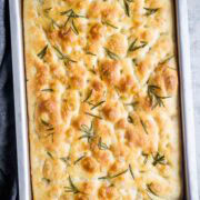 A rectangular pan of golden-brown focaccia bread, fragrant with rosemary sprigs, is held by hands using a dark cloth.