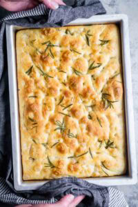 A rectangular pan of golden-brown focaccia bread, fragrant with rosemary sprigs, is held by hands using a dark cloth.