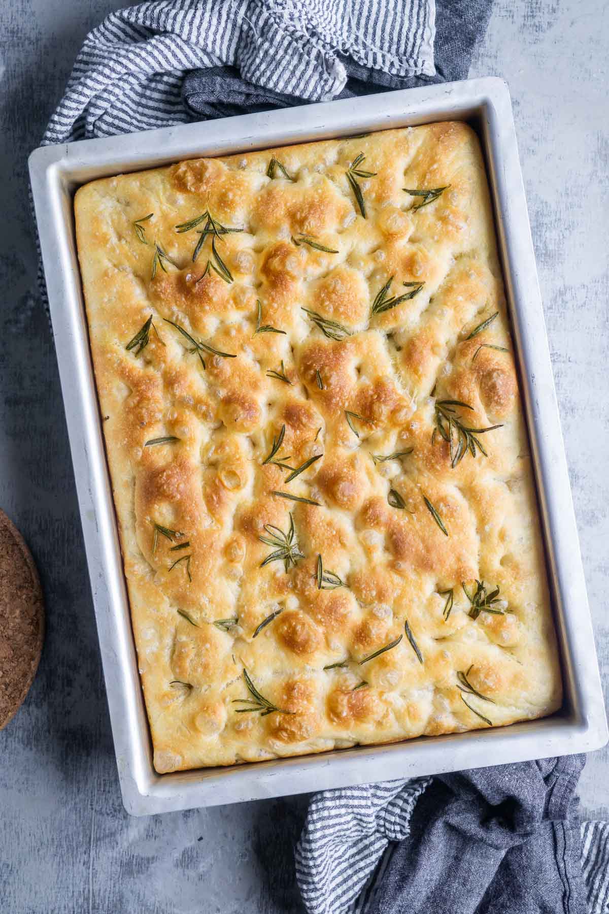 Rectangular focaccia bread in a metal baking pan, topped with rosemary sprigs and a golden, dimpled surface. A striped towel is partially visible underneath the pan, adding a cozy touch to this freshly baked focaccia bread.
