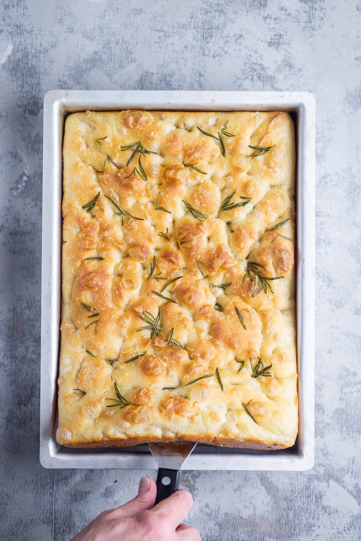 A hand holding a spatula lifts golden, freshly baked focaccia bread from a rectangular baking pan on a light gray surface.