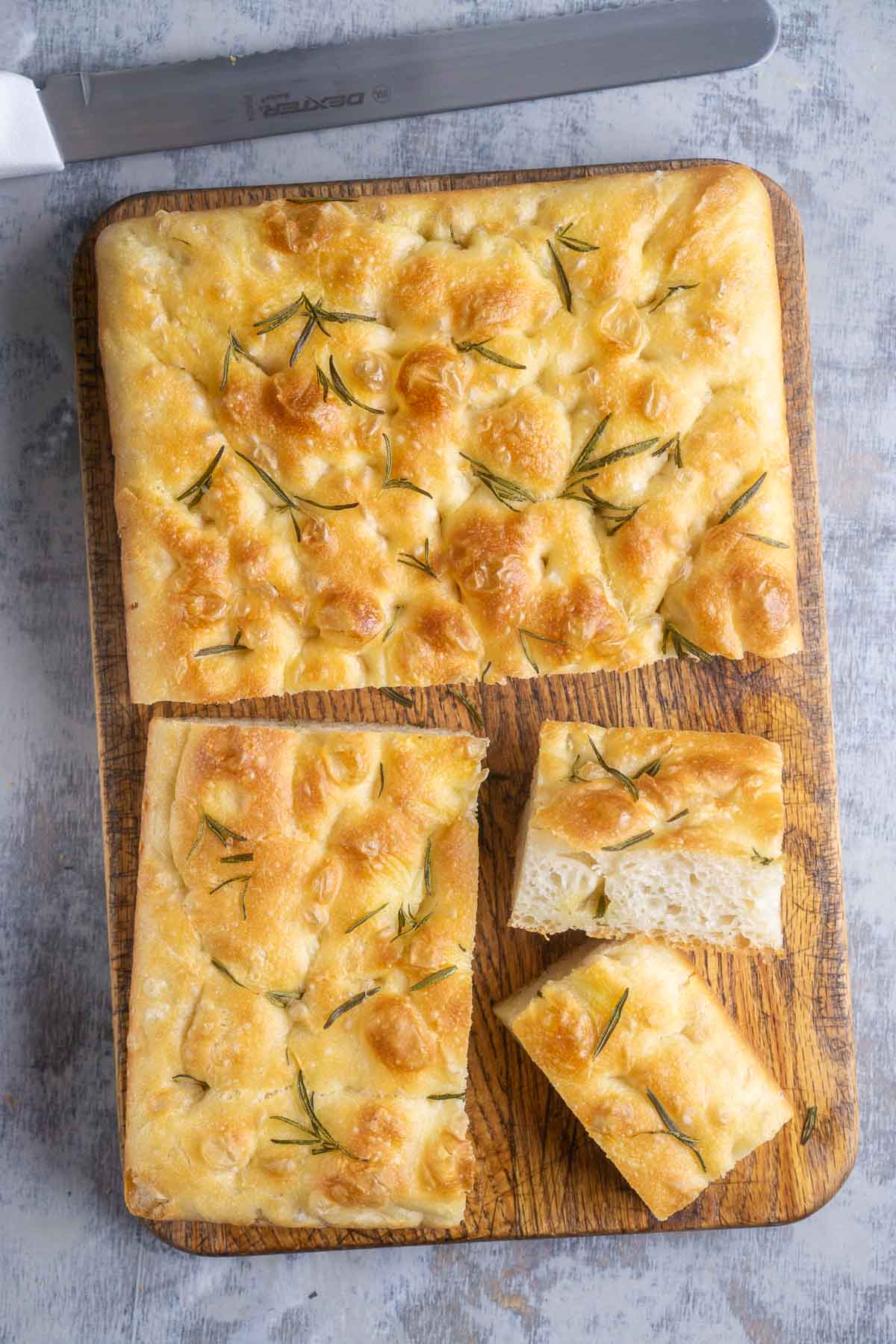 A rectangular focaccia bread topped with rosemary, partially sliced on a wooden cutting board with a bread knife nearby.
