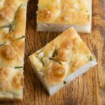 Close-up of sliced focaccia bread with a golden crust, airy interior, and sprigs of rosemary on top, displayed on a wooden board. This focaccia bread recipe showcases rustic charm and irresistible texture.